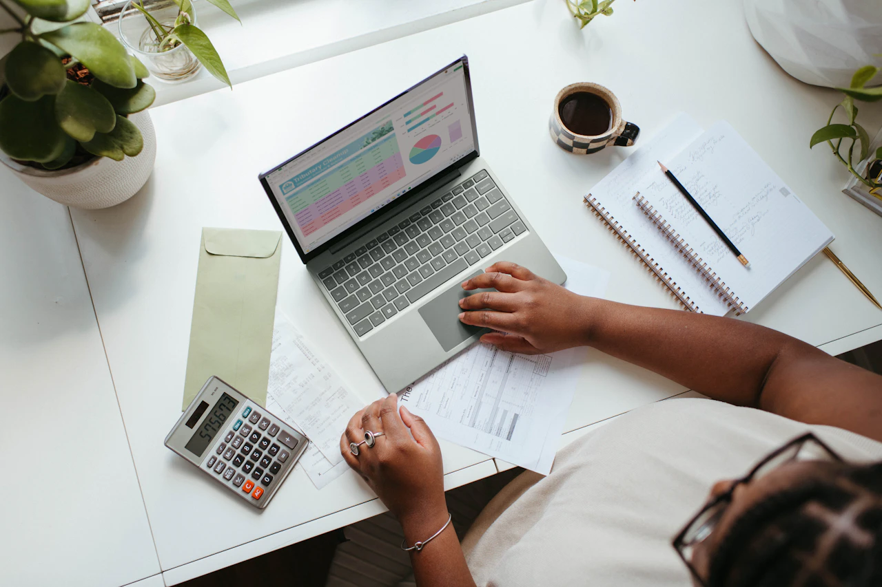 Person working with a laptop, paper records, a notebook, and a calculator